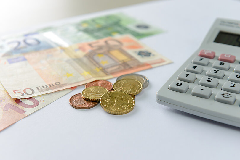 Euro-Banknoten und Münzen liegen neben einem Taschenrechner auf einem Schreibtisch.