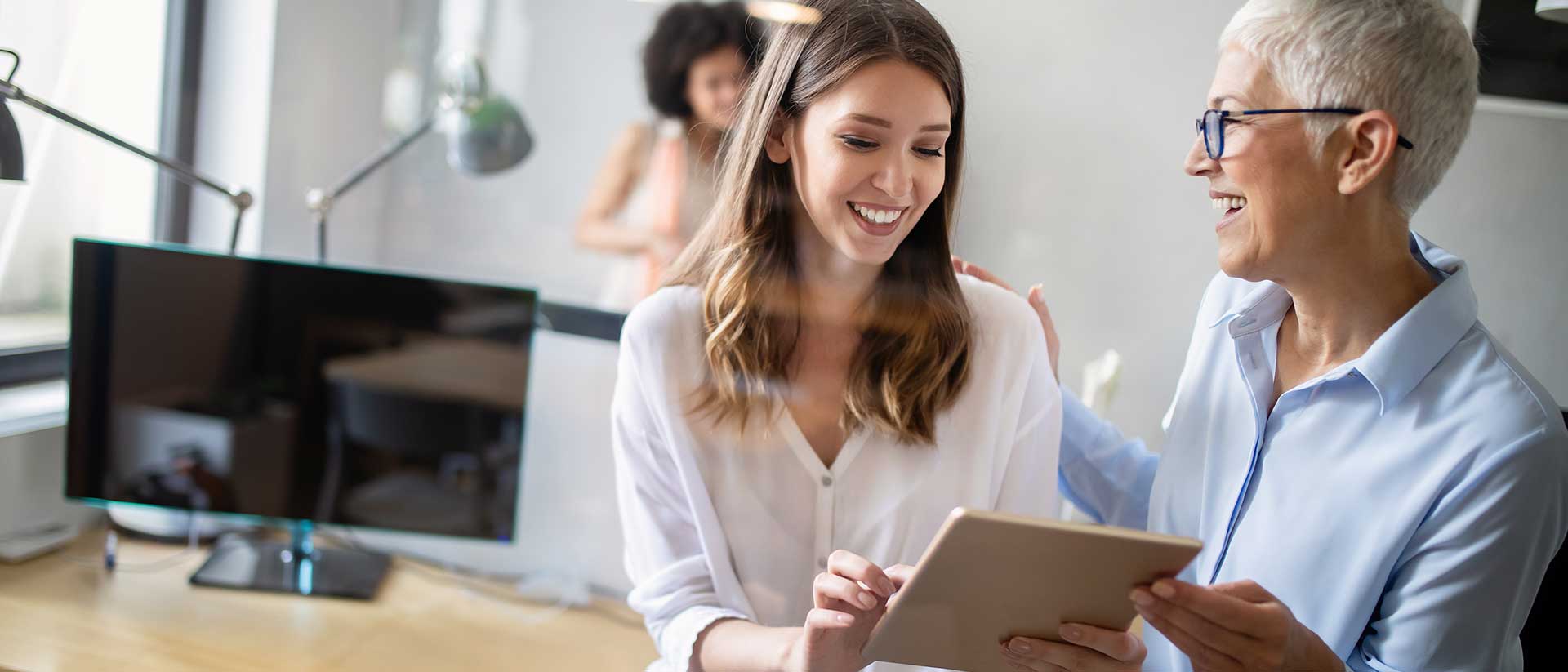 Das Bild zeigt eine junge Frau in einem Büro, die ein Tablet in der Hand hält. Im Hintergrund unterhalten sich zwei Männer.